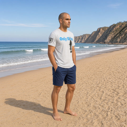 A man standing on a sandy beach wearing a white graphic t-shirt with blue text and navy blue shorts, with the ocean and rocky cliffs in the background.